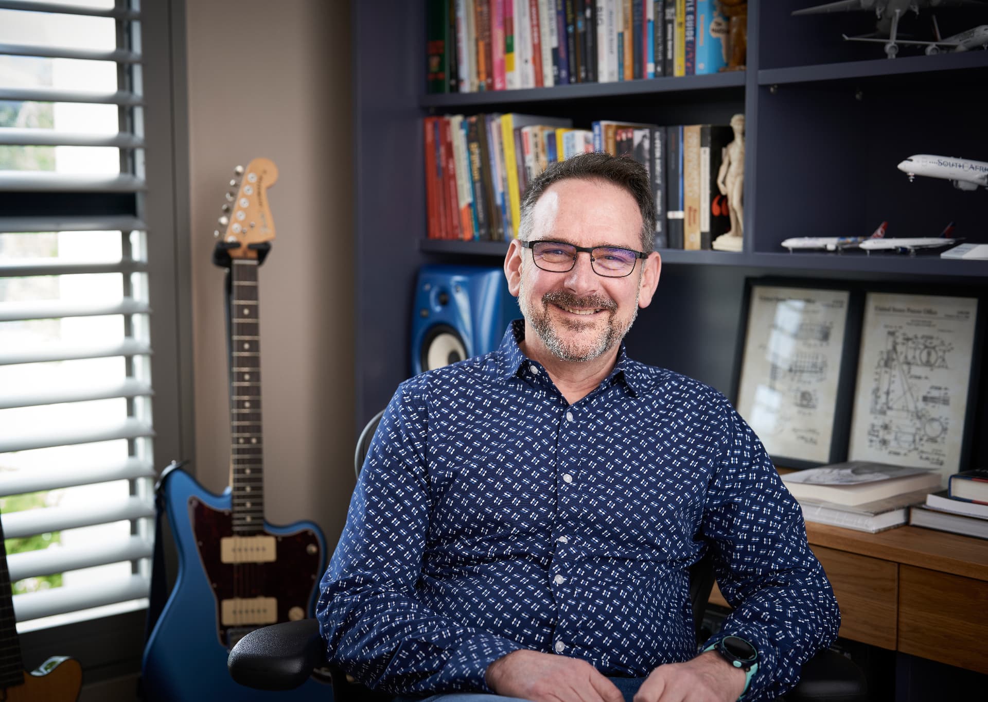 David Maclean sitting in his study, smiling, with a guitar and bookshelves behind him
